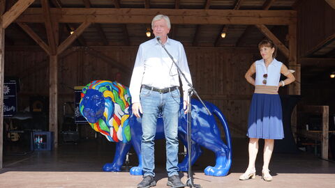 Volker Bouffier steht auf einer Holzbühne vor einer großen blau bemalten Löwen-Skulptur und hält eine Ansprache. Seine Frau Ursula Bouffier steht ein wenig abseits von ihm im Hintergrund auf der Bühne