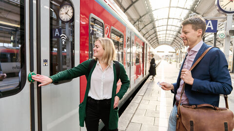 Zwei Personen stehen am Bahngleis und steigen in einen Zug ein. 