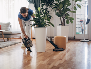 Eine Person reinigt den Holzboden in einem modernen Wohnzimmer mit einem tragbaren Staubsauger neben großen Pflanzen in weißen Töpfen.