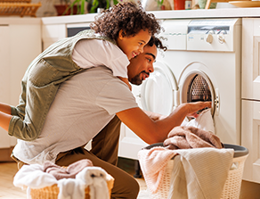 Ein Mann und ein Kind sortieren zusammen Wäsche vor einer Waschmaschine in einem gemütlichen Hauswirtschaftsraum.