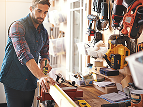 Ein Mann in einer Werkstatt misst ein Holzbrett ab. Er ist von Werkzeugen umgeben und konzentriert bei der Arbeit.