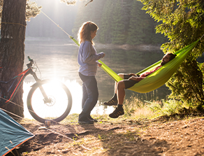 Ein Mann entspannt sich in einer Hängematte, während eine Frau daneben steht. Im Hintergrund sieht man ein Fahrrad und einen See.