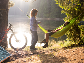 Ein Mann entspannt sich in einer Hängematte, während eine Frau daneben steht. Im Hintergrund sieht man ein Fahrrad und einen See.
