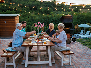 Eine Familie isst gemeinsam an einem Picknicktisch im Garten bei Abenddämmerung. Lichterketten hängen über ihnen, im Hintergrund ist ein Grill zu sehen.