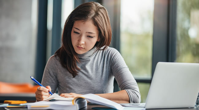 Junge Frau schreibt an einer Arbeit. Auf dem Tisch ein aufgeklappter Laptop