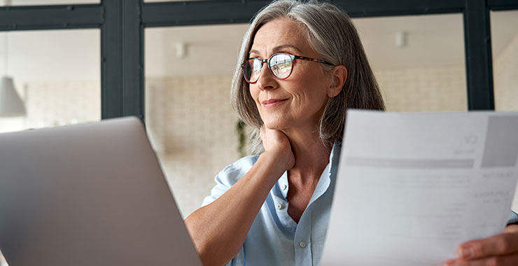 Older woman looking at laptop screen while holding resume in hand.