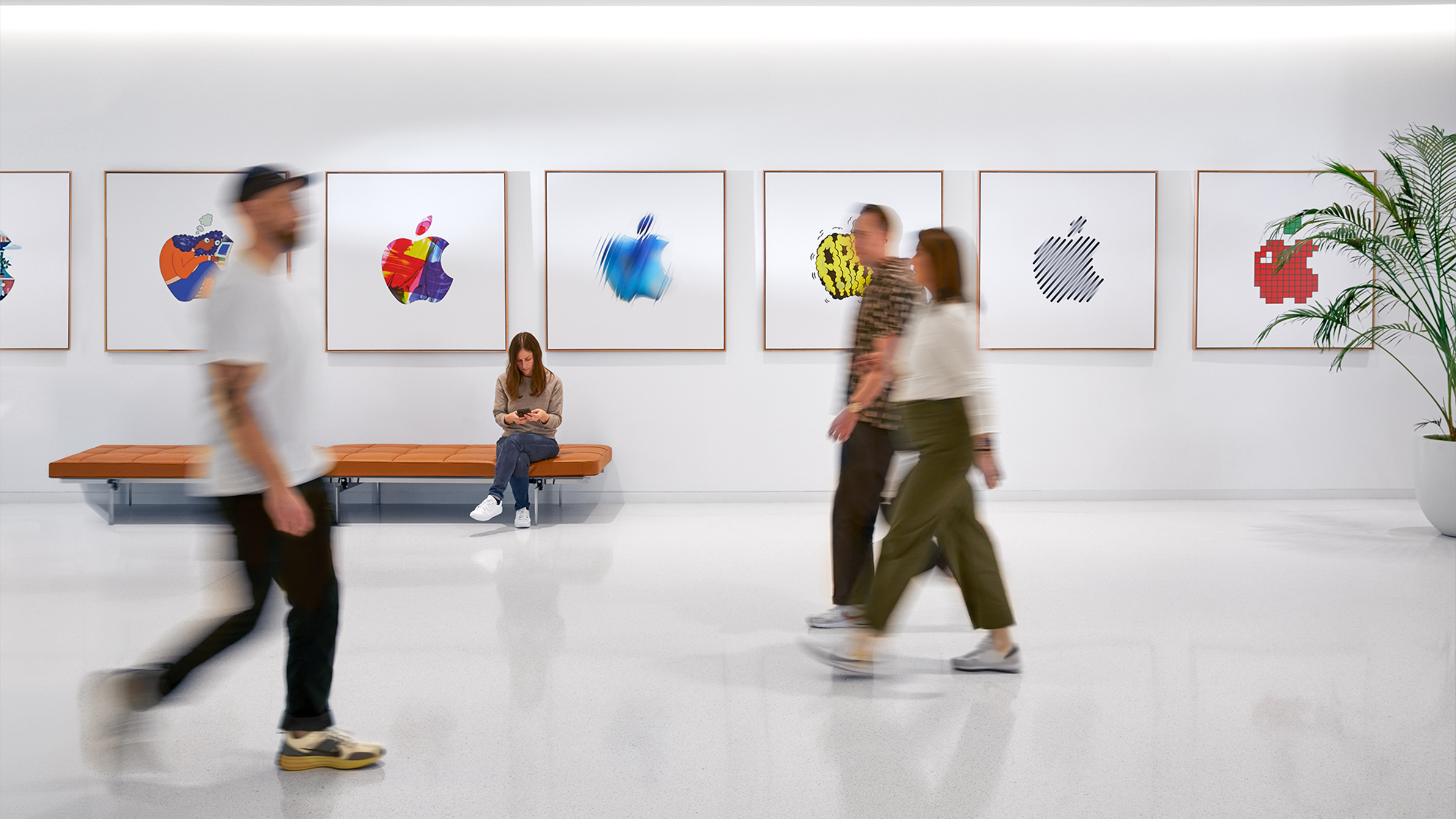 Três colegas da Apple a passar junto a uma parede com logótipos da Apple coloridos, e outra colega sentada num banco.