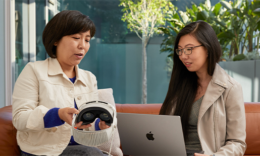 Jessica a utilizar um MacBook no interior com uma porta aberta para o pátio, sentada com uma colega da Apple, a mostrar o seu Apple Vision Pro.