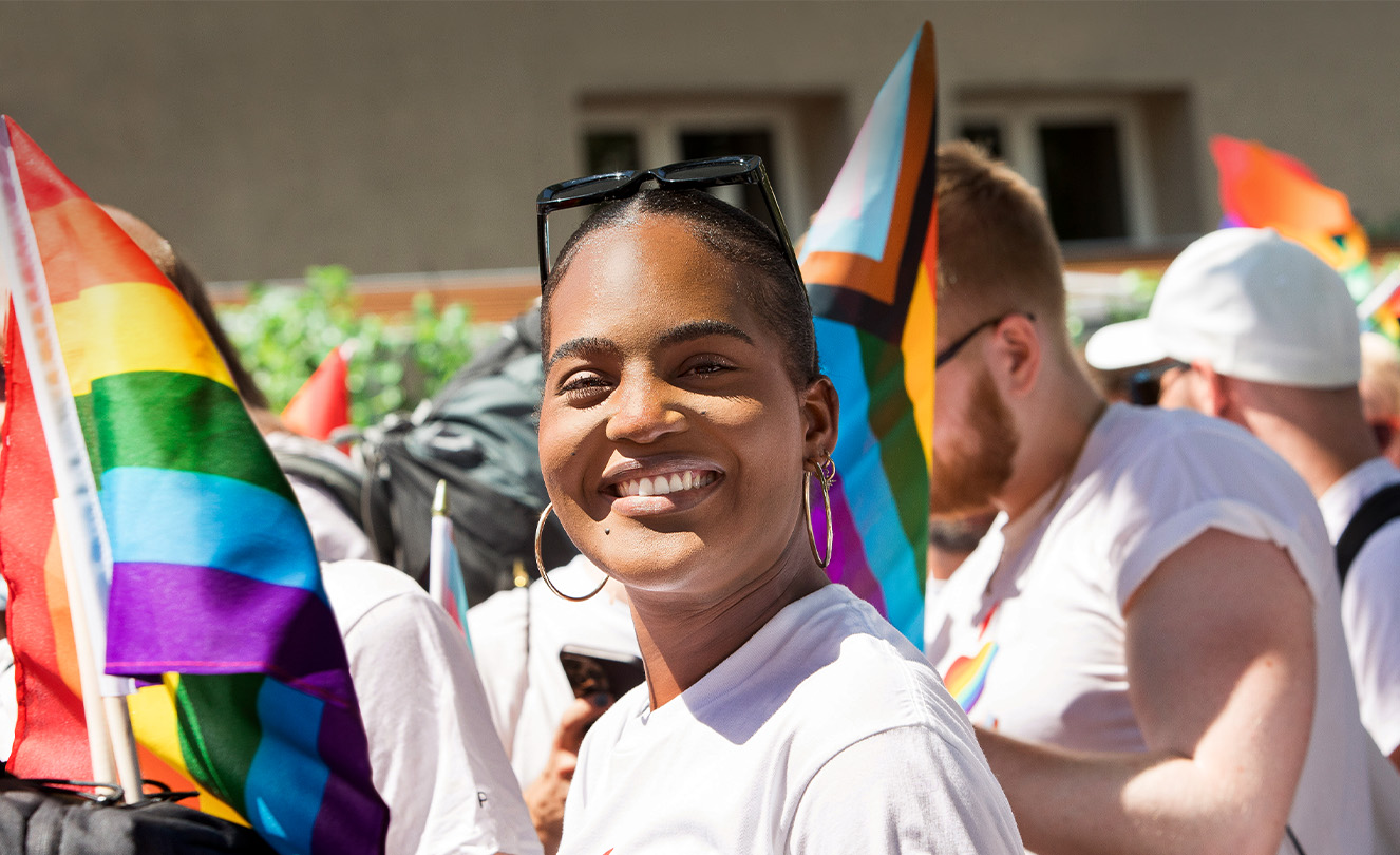 Hymyilevä Applen työntekijä Apple Pride ‑tapahtumassa, ympärillä  työntekijät Pride-t-paidoissa pitelevät Pride-lippuja. 