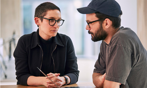 Sophie habla con un compañero que está utilizando un MacBook mientras los dos están sentados en una mesa.