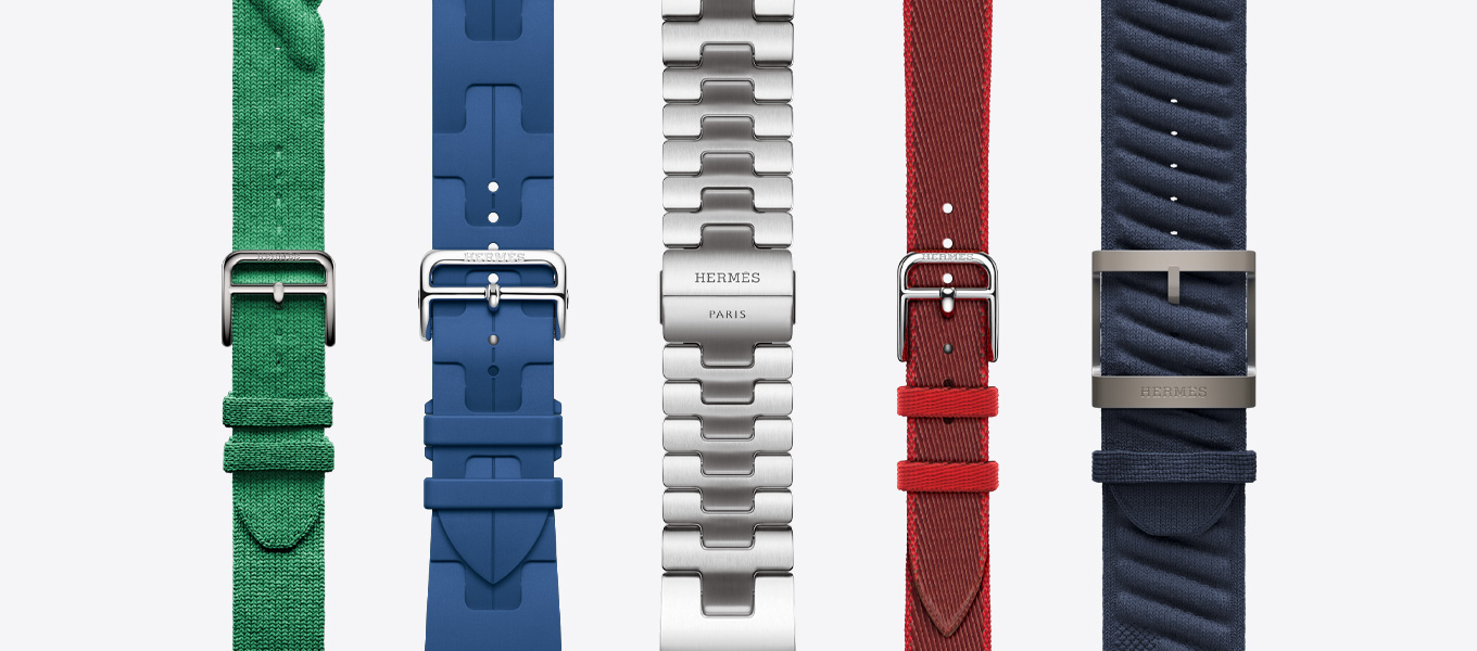 Front view of five Hermès watch bands showing a Rouge Grenat (red) Torsade, a Vert Moyen (green) Kilim, a Satiné (satin silver) Grand H, a Rouge Grenat/Vermillion (red with red trim) Twill Jump Attelage Single Tour and a Bleu Nuit (blue) En Mer.