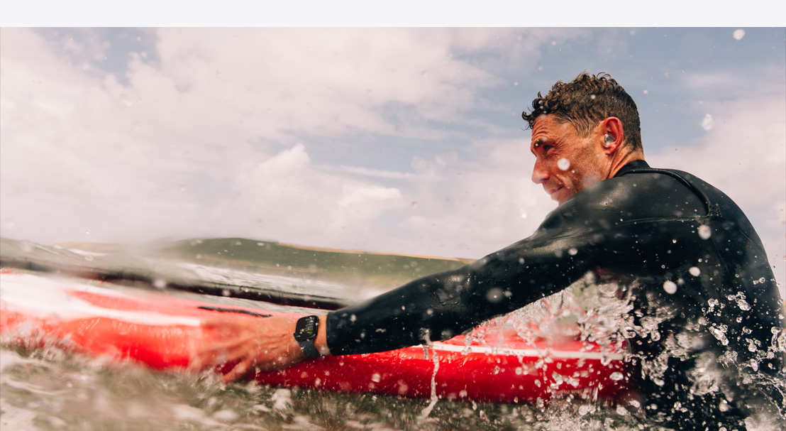 Een man in een wetsuit met een Apple Watch Series 10 om zijn pols staat in het water klaar met een surfboard.