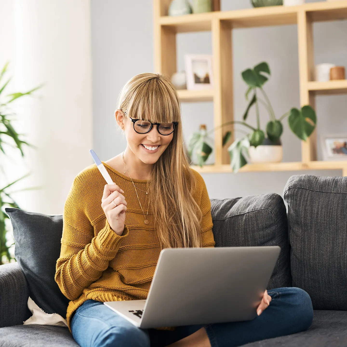 Eine Frau sitzt auf dem Sofa, guckt auf ein Laptop und hält einen Schwangerschaftstest in der Hand