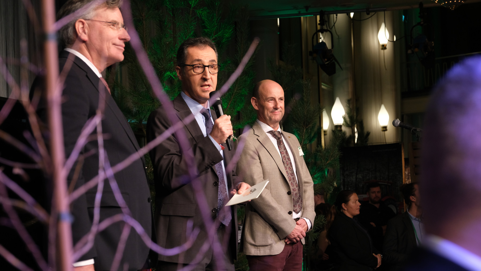 Bundesminister Cem Özdemir mit Mikrofon auf der Bühne beim Empfang der Waldbesitzer. Links und rechts von ihm stehen zwei Männer.