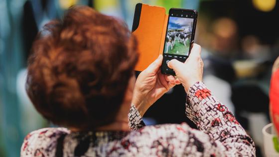 Blick über die Schulter einer Frau, auf ein Handy, mit dem sie die Messehalle fotografiert.