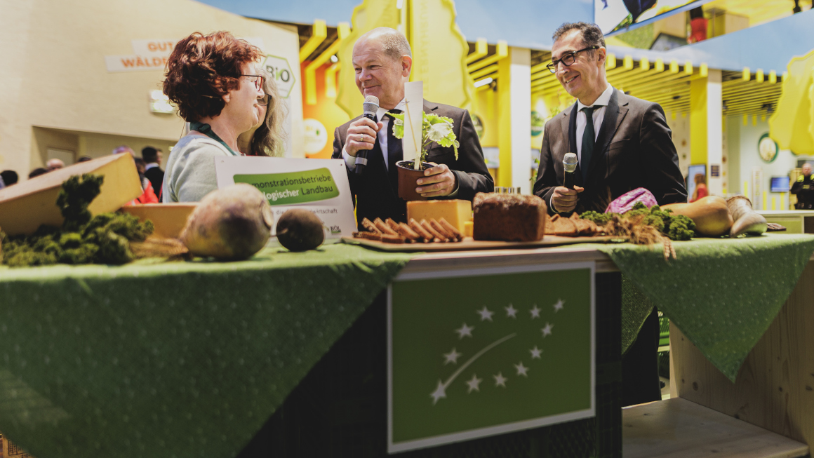 Bundeskanzler Olaf Scholz und Bundesminister Cem Özdemir stehen hinter einem Stand zum ökolgischen Landbau und informieren sich bei einer Mitarbeiterin.