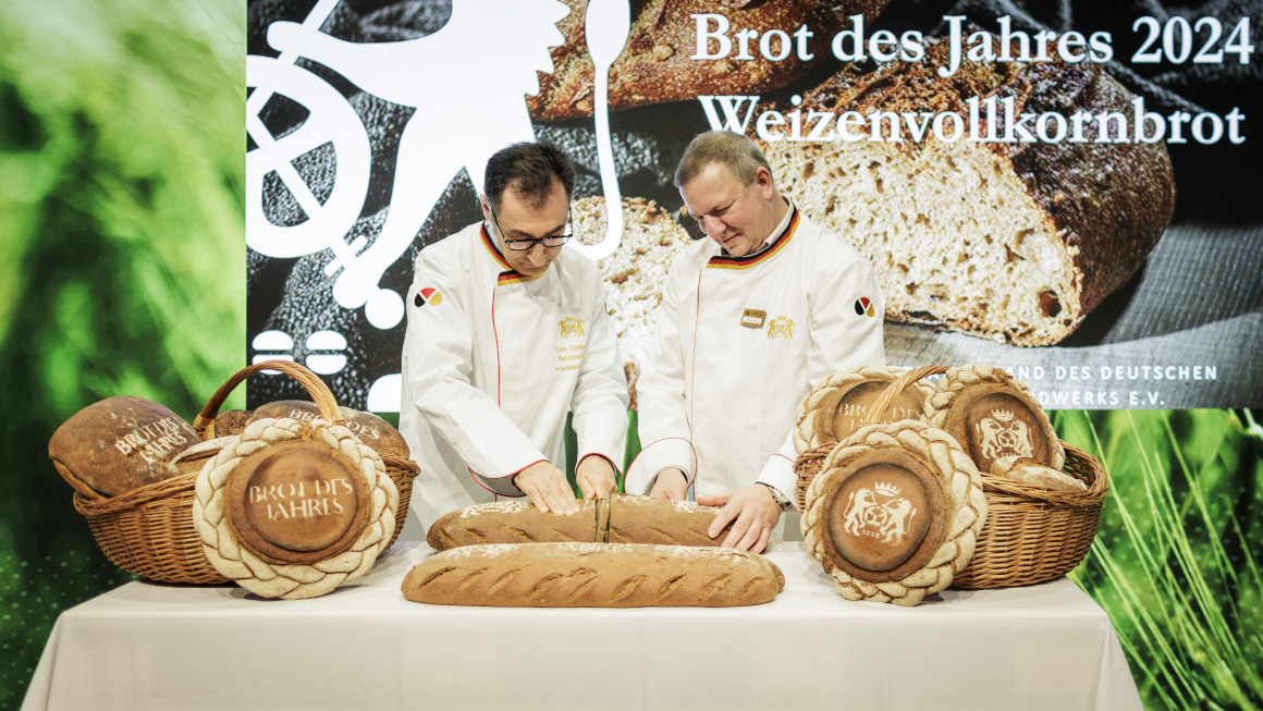 Der Bundesminister schneidet mit Roland Ermer, Präsident des Zentralverbands des Deutschen Bäckerhandwerks, ein Brot an, dahinter der Schriftzug - Brot des Jahres 2024 - Weizenvollkornbrot