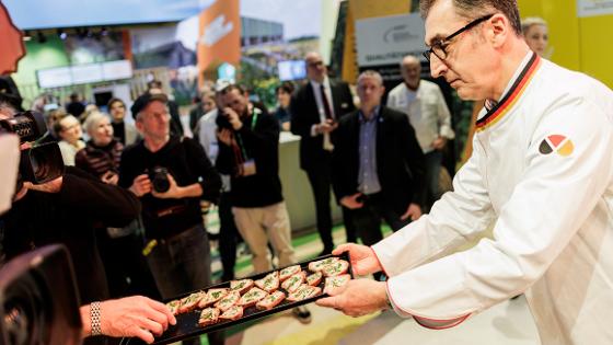 Die Bundesminister verteilt auf einem Tablett Brot mit Butter und Schnittlauch an die Hallenbesucherinnen und -Besucher.