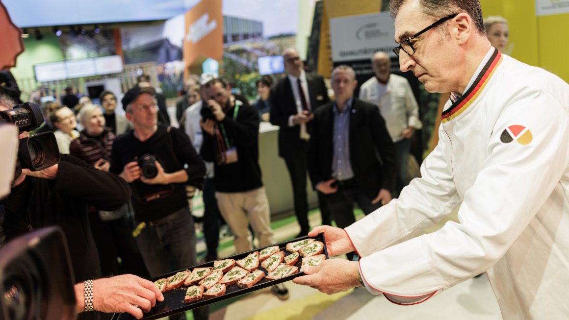 Die Bundesminister verteilt auf einem Tablett Brot mit Butter und Schnittlauch an die Hallenbesucherinnen und -Besucher.