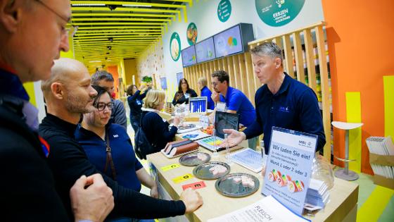 An einem Messestand unterhält sich ein Wissenschaftler mit Besucherinnen und Besuchern, diese können verschiedene Wurststücke testen.