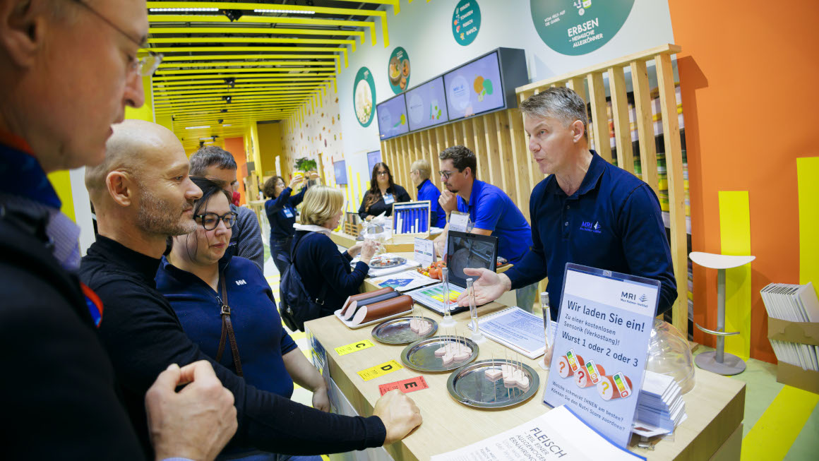 An einem Messestand unterhält sich ein Wissenschaftler mit Besucherinnen und Besuchern, diese können verschiedene Wurststücke testen.