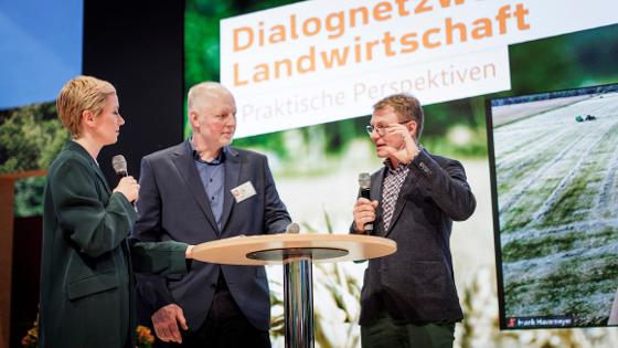 Drei Personen stehen im Gespräch auf der Bühne, dahinter der Schriftzug Dialognetzwerk Landwirtschaft - Praktische Perspektiven