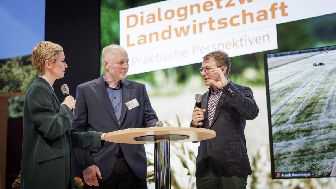 Drei Personen stehen im Gespräch auf der Bühne, dahinter der Schriftzug Dialognetzwerk Landwirtschaft - Praktische Perspektiven