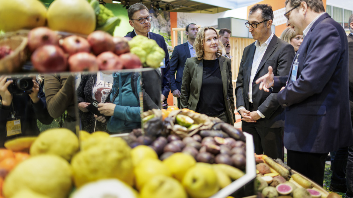 Bundesminister Özdemir und Bundesministerin Lemke stehen im Gespräch in der Messehalle an einem Gestand mit verschiedenen Obstsorten 