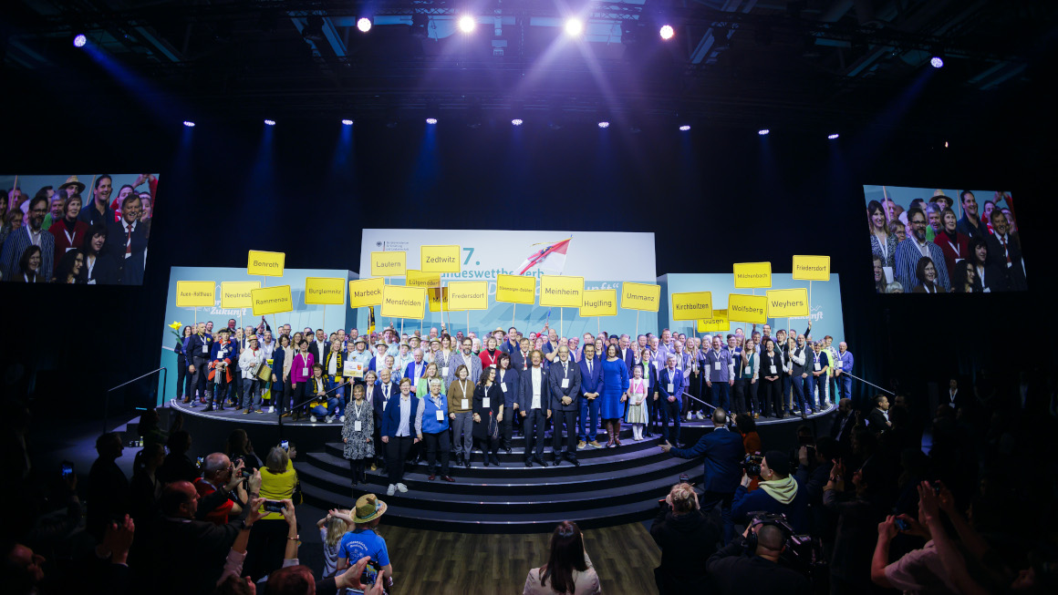 Gruppenfoto: Die Vertreterinnen und Vertreter der Siegerdörfer stehen mit Bundesminister Özdemir und der Parlamentarischen Staatssekretärin Müller auf der Bühne des Dorffestes, jedes Dorf hält ein Schild mit dem Dorfnamen in der Hand.