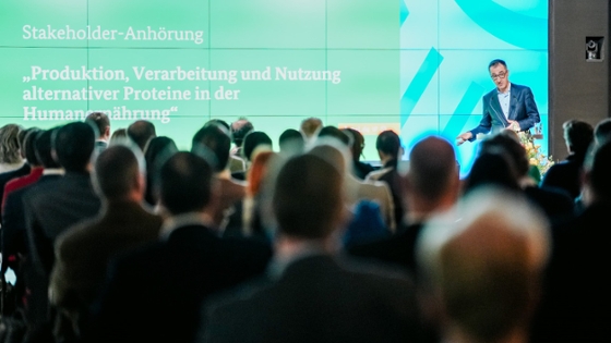 Bundesminister Cem Özdemir auf der Bühne, daneben der Schriftzug: Stakeholder-Anhörung "Produktion, Verarbeitung und Nutzung alternativer Proteine in der Humanernährung"