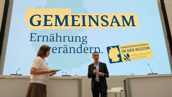 Bundesminister Cem Özdemir auf der Bühne - im Hintergrund der Schriftzug "Gemeinsam Ernährung verändern" und "Ernährungswende in der Region - Modellregionenwettbewerb"