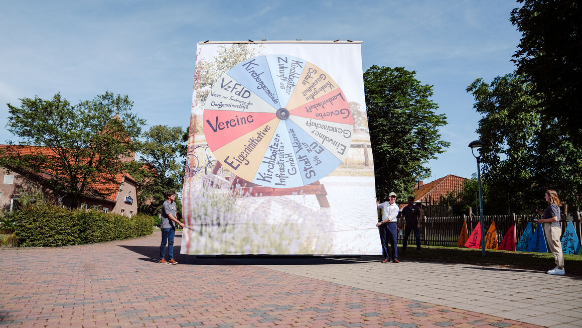 Bundesminister Özdemir steht neben einem ca. 8 Meter hohen Plakat mit einem Kreisdiagramm, von dem was Kirchboitzen ausmacht, z.B. Vereine, Eigeninitiative, Gewerbe, Kichrboitzer Zukunft e.G., Kirchgemeinde, Kita, Schule