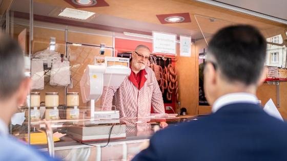 Ein Mann steht in einem Metzgereiwagen auf dem Wochenmarkt, Bundesminister Cem Özdemir ist in Rückansicht im Gespräch mit dem Mann zu sehen.