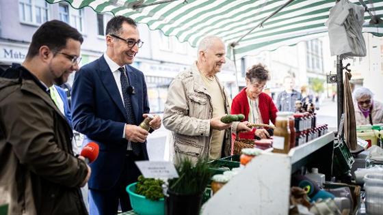 Bundesminister Cem Özdemir kauft an einem Marktstand mit Gemüse und Marmeladen ein. Neben ihm stehen weitere Marktbesucherinnen und -besucher und ein Journalist.