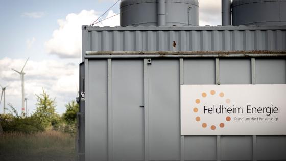 Blick auf eine Anlage  mit einem Schild, Aufschrift "Feldheim Energie - rund um die Uhr versorgt", im Hintergrund Windkraftanlange