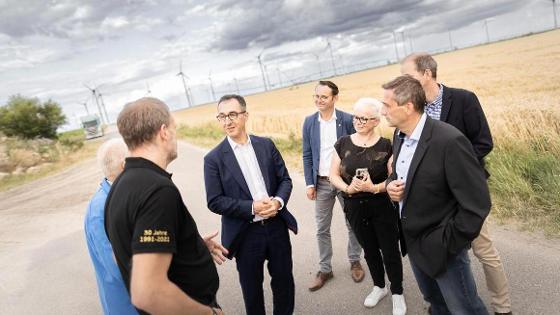 Austausch beim Besuch des Bioenergiedorfs Feldheim: Eine Personengruppe, darunter Bundesminister Cem Özdemir. Im Hintergrund ein Getreidefeld und Windkraftanlagen.