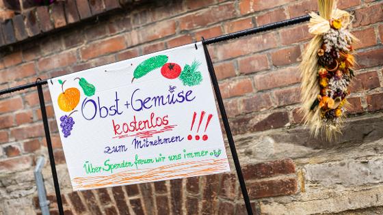 handgeschriebenes Schild mit gemaltem Obst und Gemüse und der Aufschrift "Obst+Gemüse kostenlos zum Mitnehmen! Über Spenden freuen wir uns immer" 