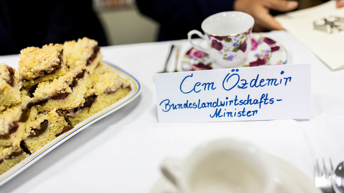 handgeschriebenes Namensschild für Bundesminister Özdemir auf einer Kaffeetafel vor einer geblümten Goldrandtasse, daneben Pflaumenkuchen