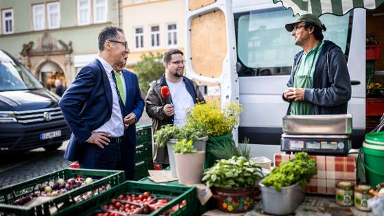 Bundesminister Özdemir auf dem Wochenmarkt im Gespräch mit einem Obst-/Gemüseverkäufer, neben ihm steht ein Radiojournalist mit Mikrofon