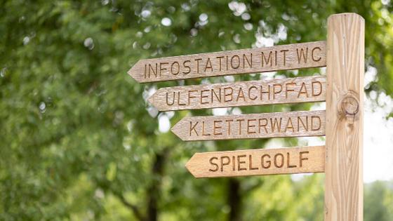 Wegweiser aus Holz mit folgenden Zielen, alle in eine Richtung: Infostation mit WC; Ulfenbachpfad; Kletterwand; Spielgolf