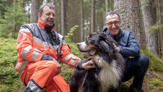 Bundesminister Özdemir und ein Rettungshundeführer knien im Wald, zwischen ihnen ein Rettungshund