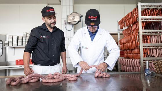 Bundesminister Özdemir probiert sich darin, Wurst abzudrehen.Neben ihm steht ein Mitarbeiter der Metzgerei.