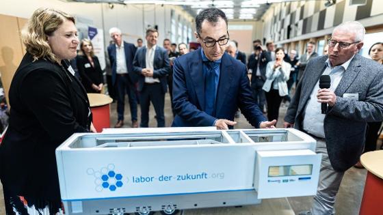 Bundesminister Cem Özdemir schaut sich das Modell eines mobilen Labors an. Neben ihm und im Hintergrund stehen weitere Personen.