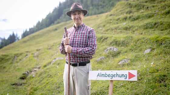 Bundesminister Özdemir steht auf der Halm mit Hut und Wanderstock, neben ihm ein Schild mit Richtungspfeil zur Almbegehung