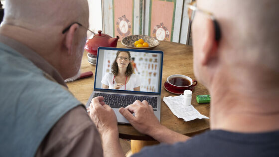 Zwei ältere Herren sitzen am Laptop und kommunizieren mit einer Ärztin, auf dem Tisch liegen Medikamente