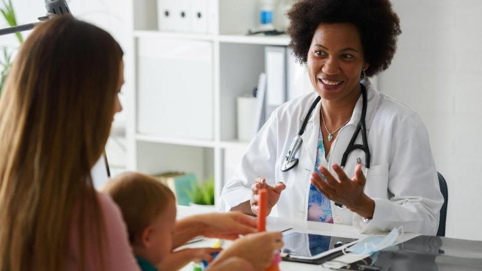 Eine Kinderärztin sitzt an ihrem Schreibtisch in einer Beratungssituation. Davor sitzt in Rückansicht eine Frau mit einem Kleinkind auf dem Schoß