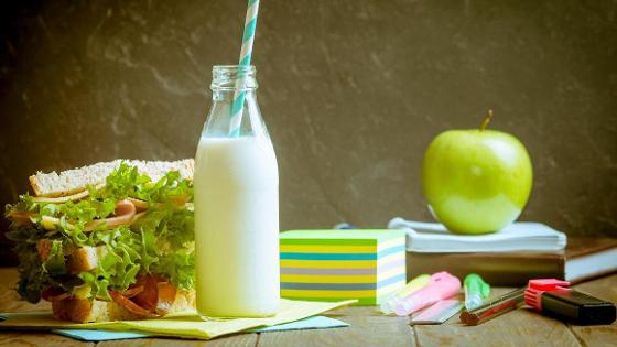 Ein belegtes Brot, eine Flasche Milch und verschiedene bunte Stifte liegen auf einem Tisch. Hinter der Flasche Milch liegen bunte Notizzettel. Im Hintergrund liegt ein Apfel auf einem Buch.