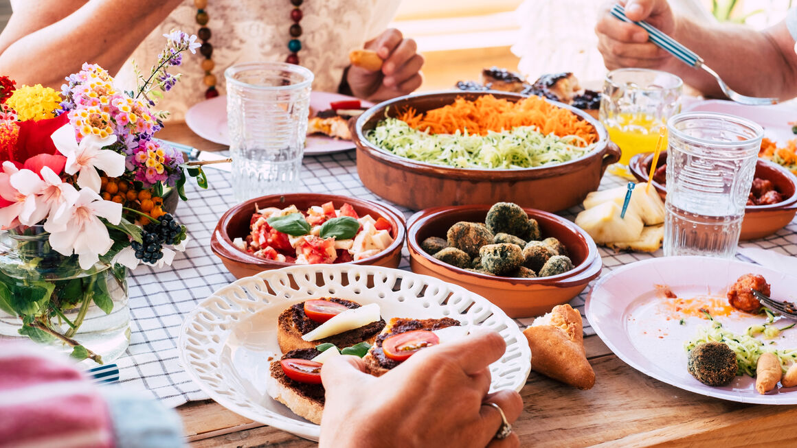 Blick von oben auf einen gedeckten Tisch mit Salat, Gemüse, Gtränken und einen bunten Blumenstrauß am Bildrand sind Personen zu erkennen