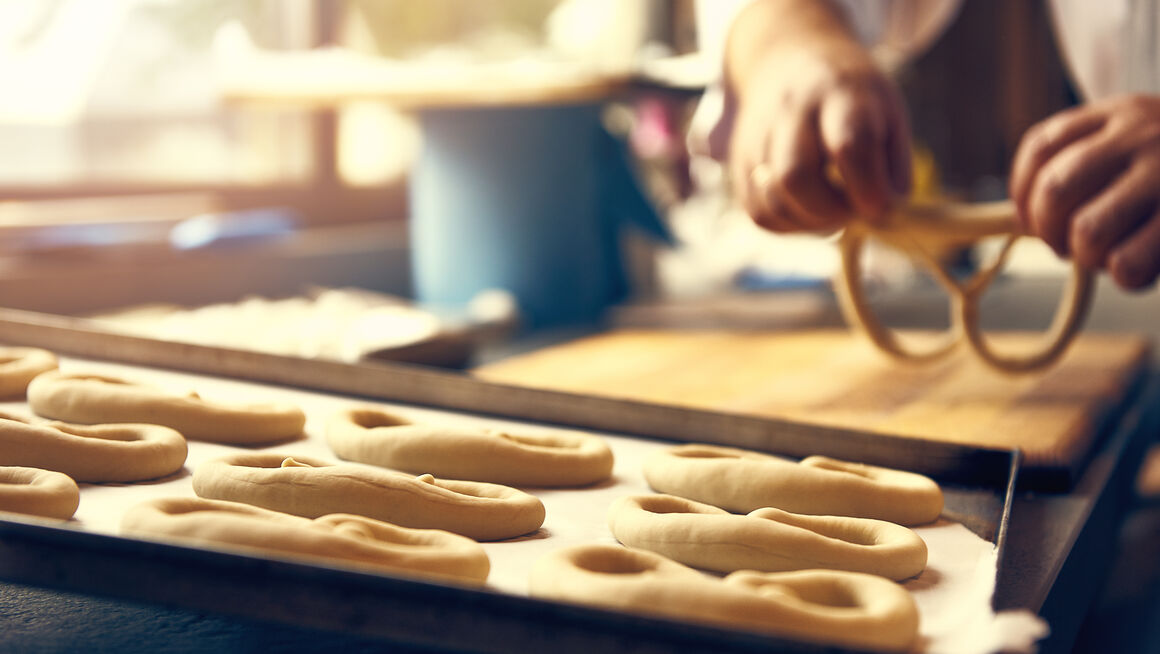 Im Vordergrund ein Blech mit Brezeln, dahinter Hände die eine Brezel formen