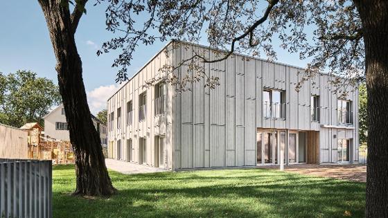 Außenansicht eines Gebäudes aus hellem Holz, im Vordergrund Rasen und Bäume, neben dem Gebäude ein Holz-Spielplatz